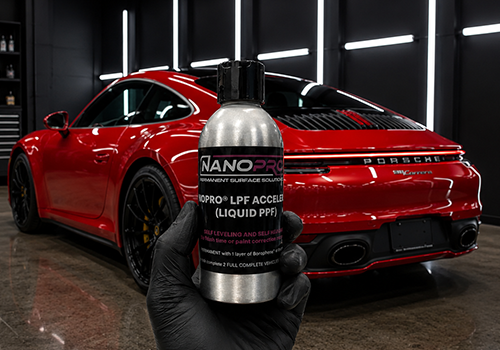 Gloved hand holding a silver bottle labeled Nanopro LPF Accelerator in a car detailing workshop with a red Porsche in the background.